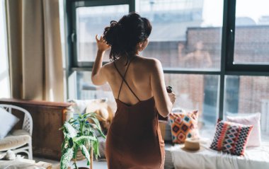 Terracotta elbiseli, tanınmayan esmer, ayakta duran, saçlarına dokunan, pencereden dışarı bakan ve modern aydınlık dairede tefekkür eden...