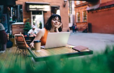 Siyah saçlı genç bir kadın, tahta masada oturuyor ve uzaktan kumandalı bir proje üzerinde çalışırken, sokak kafesinde dizüstü bilgisayar kullanıyor.