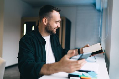 Yapışkan notlar ve kağıtlarla masada oturan olumlu erkek girişimcinin yan görünümü planlayıcıyı kontrol ederken iş yerinde akıllı telefona göz atıyor