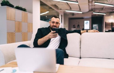 Genç sakallı erkek boş zamanlarında bilgisayar ve cep telefonuyla masanın yanındaki koltukta dinleniyor. Ekrana bakıyor ve ofisteki modern çalışma alanında proje üzerinde çalışıyor.