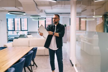 Siyah ceketli ve gözlüklü sakallı bir erkek çalışanın kollarını sıvayıp iş yerinde çalışırken bilgisayar tutarken başka yöne bakıyor.