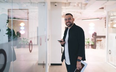 Gözlüklü, güler yüzlü erkek girişimci modern çalışma alanında elinde not defteriyle duruyor ve ofiste buluşmaya hazırlanırken cep telefonuyla kameraya bakıyor.