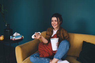 Siyah saçlı, gülümseyen genç bir kadın bacak bacak üstüne atmış koltukta oturuyor ve elinde patlamış mısırla uzaktan kumandayla kanal değiştiriyor.