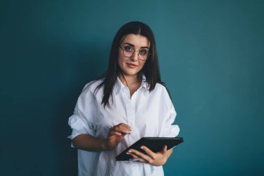 Gözlüklü, beyaz tişörtlü, tablet kullanan ve koyu mavi arka planda kameraya bakan modern bir esmer.