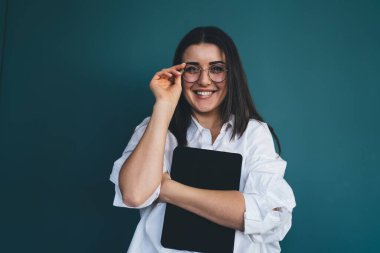 Şık beyaz gömlekli, şık gözlükler takan ve mavi arka plandaki kameraya neşeyle bakarken tableti kucaklayan pozitif genç bir kadın.