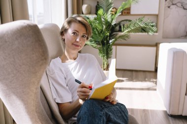 Açık renk saçlı bayan boş zamanlarında koltukta oturup not defterine not alırken kameraya bakıyor.