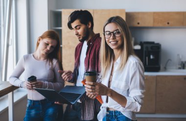 Bir grup çok ırklı ofis çalışanı bir fincan kahve ve taşınabilir bilgisayarla ofis mutfağında iş birliği yapıyor.
