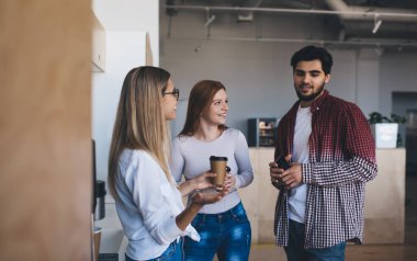 Bir grup mutlu çoklu etnik iş arkadaşı gündelik giysiler içinde modern ofiste kahve molası sırasında sıcak içecekler ve iletişim kuruyorlar.