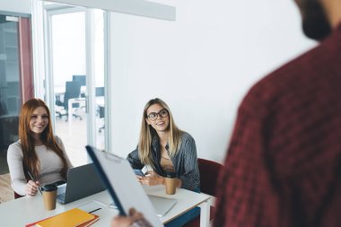 Masada dizüstü bilgisayarlı kahve fincanları ve akıllı telefonla oturan genç ve pozitif bayan çalışanlar. Ve elinde bir not panosuyla projeye katılan isimsiz erkek meslektaşlarının derslerine katılırken kameraya bakıyorlar.