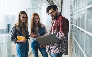 Ellerinde not defteri olan genç, gülümseyen kadınlar dizüstü bilgisayarlı erkek meslektaşlarıyla birlikte ekranlara bakarken aynı zamanda dış cam ve ofis duvarları ile etkileşime geçip zaman geçiriyorlar.