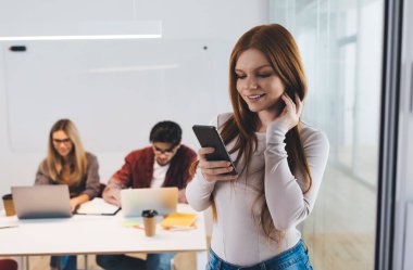 Şık giyinen gülümseyen kadın çalışan dizüstü bilgisayarlı iş arkadaşlarına karşı başarılı bir proje üzerinde çalışırken cep telefonunda okuma ve mesaj yazma takınıyor.