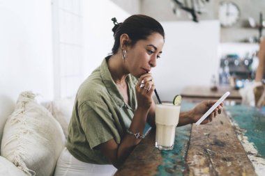 Latin asıllı genç bir kadının günlük kıyafetleriyle ahşap masada milkshake ile oturması ve cep telefonu ekranına bakması.