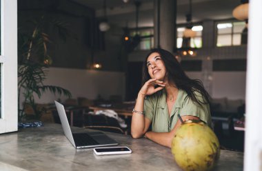 Rahat kafeterya projesinde çalışırken laptoplu akıllı telefon ve hindistan cevizli masada oturan ve gözlerini kaçıran gündelik kıyafetli genç bir kadın.