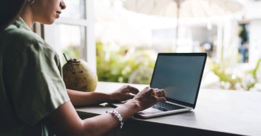 Günlük giysiler içinde serbest çalışan bir kadın netbook ile masada oturuyor ve samanla taze hindistan cevizi yerken uzak bir proje üzerinde çalışıyor.