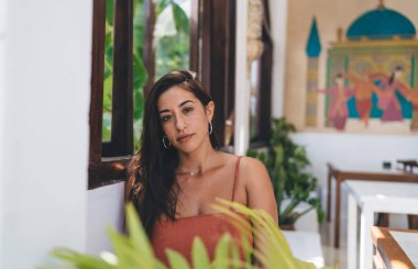 Uzun siyah saçlı, düşünceli genç bir kadın. Günlük kıyafetleriyle duvara yaslanıyor ve doğu evindeki saksı bitkileriyle süslenmiş kameraya bakıyor.