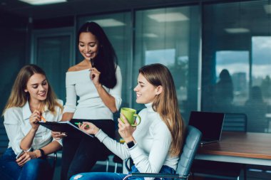 Gündelik giysiler içinde, masa başında not panosu ve dizüstü bilgisayarı olan, modern çalışma alanındaki toplantı sırasında iş projesinden bahseden bir grup gülümseyen çok ırklı genç kadın.