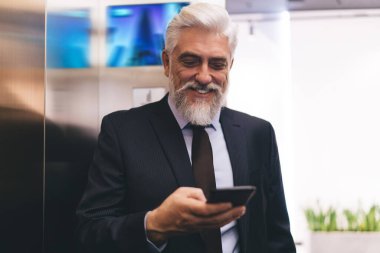 Zarif takım elbiseli, gülümseyen olgun iş adamı modern ofiste duvarın yanında dururken ekrana ve cep telefonuna bakıyor.