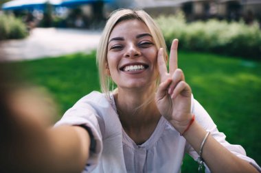 Ziyaretçi işareti gösteren ve gülümseyen neşeli bir kadının selfie portresini kapatın. Seyahat yaşam tarzından zevk alan mutlu beyaz kadın, içeriği etkilediği için boş zamanlarında kameraya gülüyor.