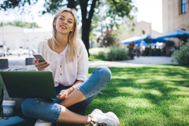 Web tasarımı projesinde uzaktan çalışmak için kameraya gülümseyen mutlu kadın serbest yazar portresi, kampüs parkında cep telefonu ve dizüstü bilgisayarla poz veren neşeli beyaz kadın.