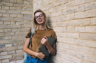 Kafkasyalı dijital göçebenin yarı boy portresi görüş düzeltmesi için optik gözlüklerle duvarın yakınındaki kameraya bakıyor, milenyum kadın blogcusu şehirde kapalı dizüstü bilgisayar teknolojisiyle poz veriyor