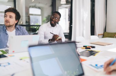 Modern ofis projesindeki toplantıya katılırken dizüstü bilgisayarlı belgeler ve meçhul iş arkadaşlarıyla masaya otururken başka tarafa bakan bir grup konsantre çok ırklı meslektaşım.