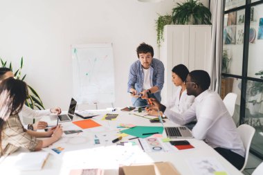 Proje ve erkek sunucu elinde kalem ve yapışkan notla kameraya bakarken masada kağıt ve dizüstü bilgisayarla oturan çok ırklı meslektaşlar.