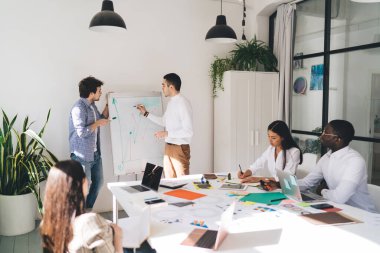 Erkek meslektaşlar yeni iş planlarını çizerken aynı zamanda çok ırklı iş arkadaşlarına dizüstü bilgisayarlar ve kağıtlarla masaya oturarak notlar alıp dinliyorlar.