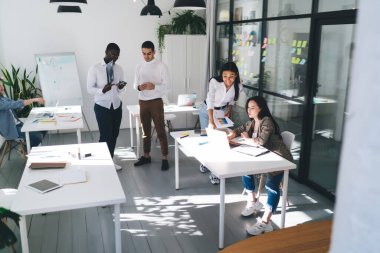 Yoğunlaşmış kadın meslektaşlar masada toplanıyor ve modern çalışma alanında ayakta duran ve akıllı telefonları karıştıran çok ırklı iş arkadaşlarının ekranlarına bakıyorlar.