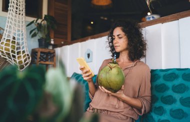 Sosyal medyada sohbet etmek ve mesajlaşmak için cep telefonu aygıtı kullanan, tropik içeceği olan beyaz bir kadın, modern akıllı telefonlu, internet sitesi için 4G kablosuz ağa bağlanan bin yıllık bir kadın.
