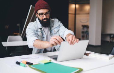 Yoğunlaşmış sakallı, günlük giysiler içinde bir erkek işçi dosya kâğıdı not defteriyle masada oturuyor ve ekranlara bakarken ve modern çalışma alanında çalışırken dizüstü bilgisayarı elleriyle temizliyor.
