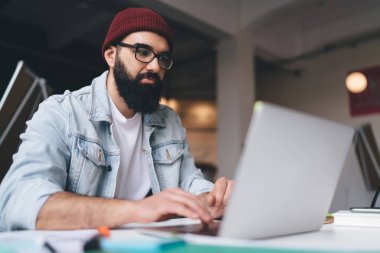 Ciddi sakallı, akıllı giysiler ve gözlüklü serbest çalışan bir erkek dizüstü bilgisayar ve yaratıcı iş yerinde proje üzerinde çalışırken masada oturuyor.