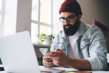Akıllı elbiseli ve gözlüklü, odaklanmış sakallı bir erkek yönetici. Masada dizüstü bilgisayarla oturuyor ve ofisteki projede çalışırken akıllı telefonlara bakıyor.