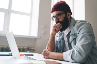 Ofisteki pencerenin yanındaki dizüstü bilgisayarda çalışırken masasında oturan şık elbiseli ve gözlüklü serbest çalışan sakallı bir erkek.
