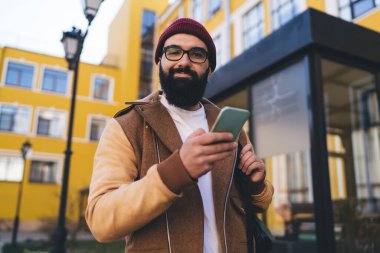 Sırt çantası sokakta duran ve cep telefonunu bulanık arka planda tutarken kameraya bakan sıradan giysili genç sakallı adamın alçak açısı.