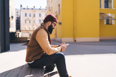 Sokak tarzı giyinmiş, gözlüklü sakallı bir adam sosyal ağlarda konuşurken dinlenirken izinli günlerinde cep telefonu kullanıyor.