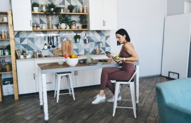 Modern mutfakta masada otururken avokado ve elma arasında seçim yapan taşınabilir bilgisayarı olan kafası karışmış bir kadın.