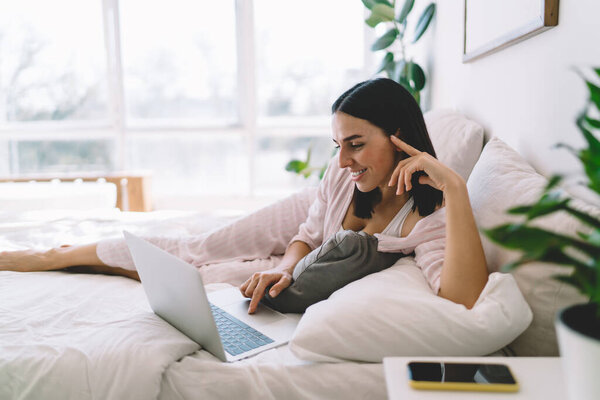Вид сбоку счастливой женщины в сонной одежде, серфингующей по Интернету на компьютере, и охлаждающей дома в современной спальне по выходным