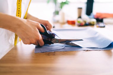 Elinde mezurası olan isimsiz bir kadın terzi ahşap masada duruyor ve elinde bulanık bir atölye ile mavi renkli kumaşı kesiyor.