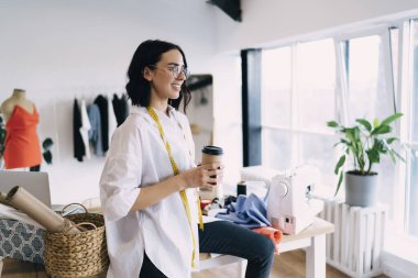 Gözlüklü, mutlu, genç bayan terzinin dikiş makinesiyle masaya oturması ve kahve içerken gözlerini kaçırması.