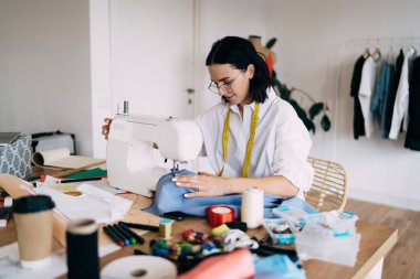 Gözlüklü genç bir kadın stüdyoda masada oturuyor ve iş günü kumaş dikerken konsantre oluyor.