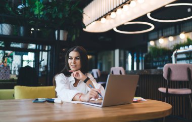 Siyah kayışlı saati olan, elinde kalemle ahşap masada dizüstü bilgisayarlı akıllı telefon kırtasiyesiyle oturan ve kafede işe başlarken gözlerini kaçıran genç bir kadın.