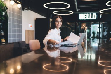 Masasında dizüstü bilgisayar ve kağıt belgelerle oturan, modern açık uzay ofisinde çalışan ve kameraya bakan beyaz tişörtlü genç bir esmer.