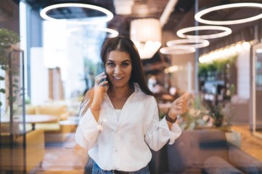 Aydınlatıcı ışıklarda duran ve modern kafede kameraya bakıp cep telefonuyla konuşan pozitif genç bir kadın.