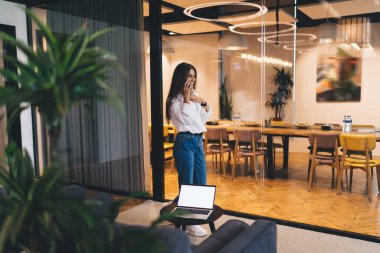 Mutlu, serbest çalışan, günlük giysiler giyen, cep telefonuyla konuşan, netbook 'la masanın yanında duran ve çalışırken gülümseyen bir kadın.