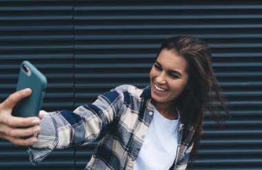 Uzun siyah saçlı, kareli tişörtlü neşeli, kendine güvenen genç bir kadın. Caddedeki metal duvarın yanında dururken gülümsüyor ve akıllı telefondan selfie çekiyor.