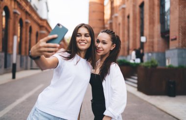 Gülümseyen genç bayan arkadaşlar eski şehrin kaldırımlarında durup akıllı telefon kameralarına bakarken kendi fotoğraflarını çekiyorlar.