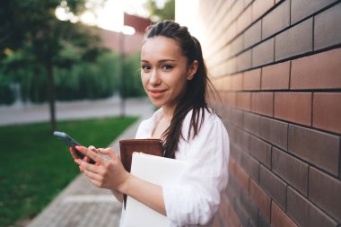 Beyaz tişörtlü pozitif genç kız kameraya bakarken tuğla duvarın yanında duruyor ve cep telefonuyla mesajlaşıyor.