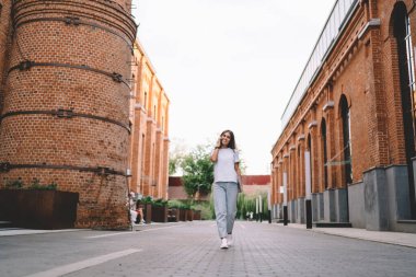 Düşük açılı, neşeli, günlük kıyafetli, dizüstü bilgisayarlı bir kadın yazın akıllı telefondan konuşurken şehir caddesinde yürüyor.