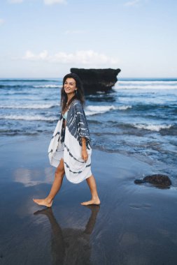 Yalınayak gezen, yaz kıyafetleri içinde uzun siyah saçlı, ıslak kumlu okyanus sahillerinde yürürken gülümseyen ve kameraya bakan genç kadın yolcuların yan görüntüsü.