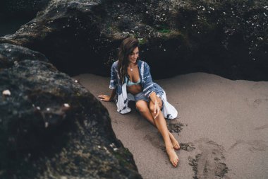 Yüksek açılı, çıplak ayaklı genç bir kadın çıplak ayakla kumlu sahilde dinleniyor ve kayalık oluşumlara bakıyor.